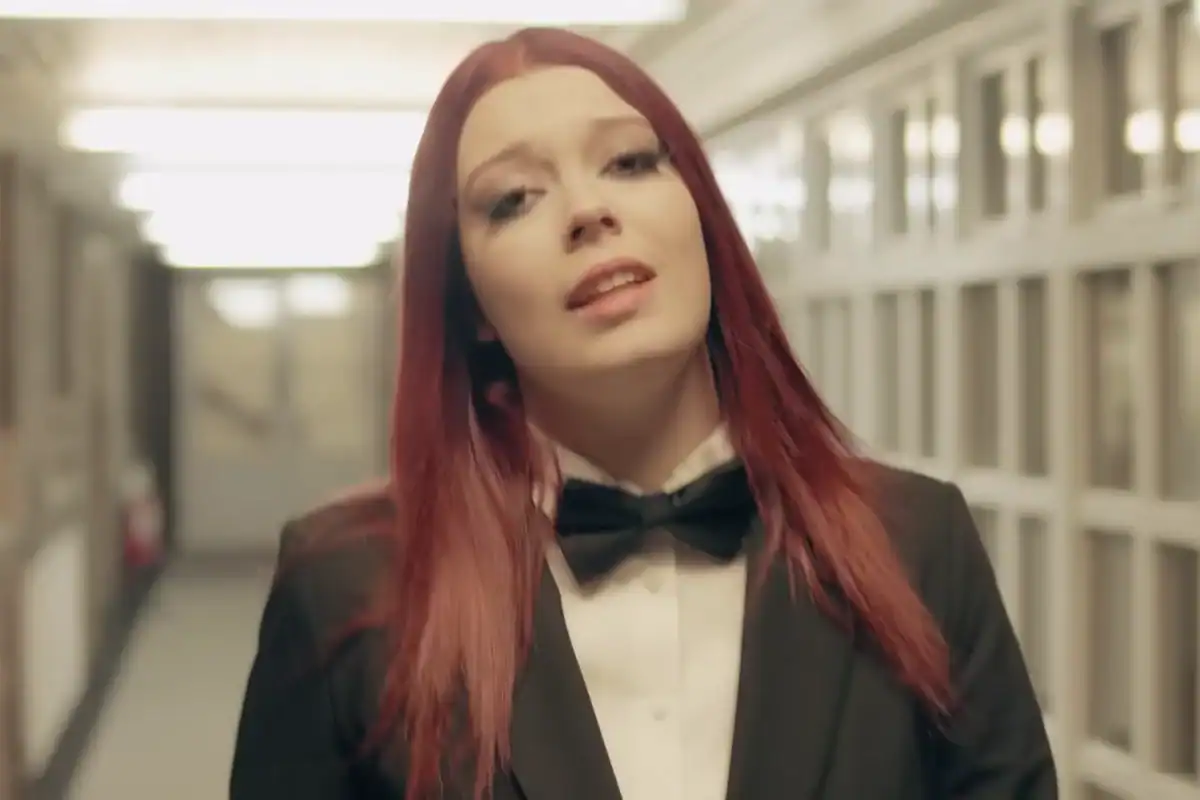 Female singer with red hair in a black tuxedo and bow tie, performing in a modern indoor setting.